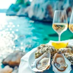 Croatian food. Fresh Oysters on white plate with lemon and and two glasses of white wine on blurred seascape background. Healthy seafood. Fresh Oyster in open air restaurant. Gourmet food. Copy space